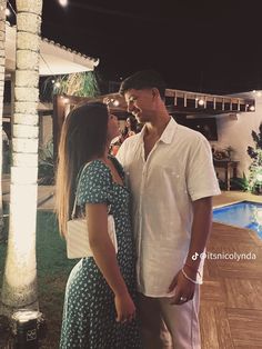 a man and woman standing next to each other in front of a pool at night