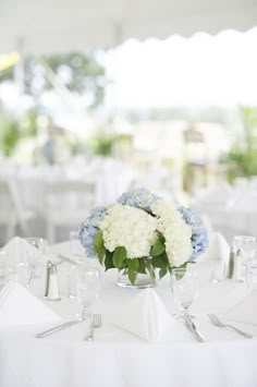 white and blue flowers are in a glass vase on the dining table set for an event