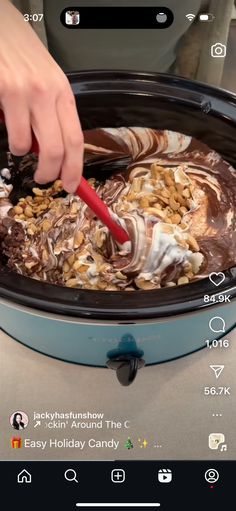 someone using a red spatula to dip some food into a crock pot filled with chocolate and marshmallows