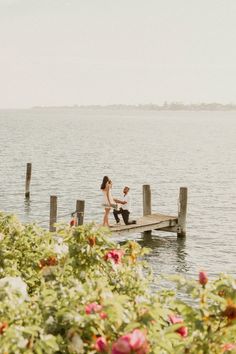 a man and woman sitting on a dock near the water with flowers in front of them