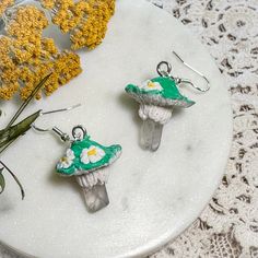 two green and white earrings sitting on top of a table next to yellow dandelions