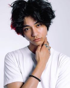 a man with black hair wearing a white t - shirt and holding his hand on his chin