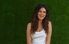 a woman standing in front of a green grass wall with her hands together and looking at the camera