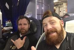 two men sitting next to each other on an airplane, one giving the peace sign