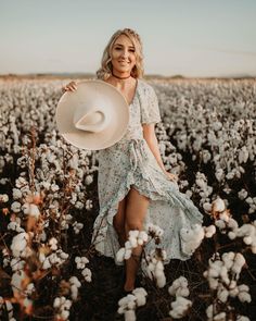 a woman standing in a cotton field holding a white hat and smiling at the camera