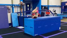 a woman is doing an acrobatic trick on a blue box in a gym