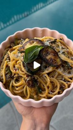 a person holding a bowl of pasta with mushrooms and spinach on the table next to a swimming pool