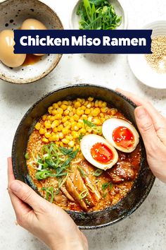 two hands holding a bowl of food with eggs, corn and other foods on the table