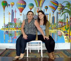 two women sitting on a white chair in front of a wall with hot air balloons