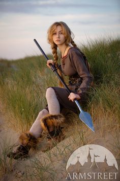 a woman sitting in the sand with two swords and a long tail on her knee