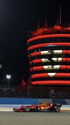 a red race car driving down a track in front of a large building at night