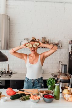 a woman standing in front of a kitchen counter holding two slices of avocado over her eyes