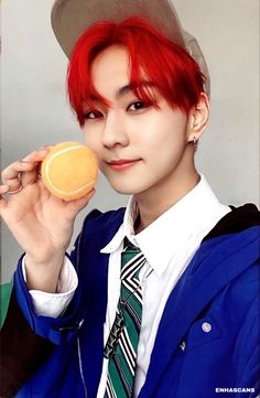 a young man with red hair is holding an orange and looking at the camera while wearing a hat