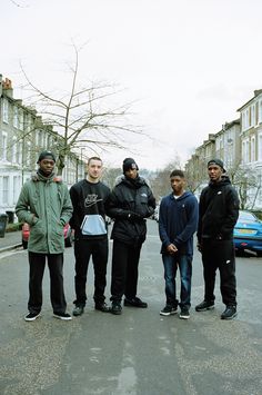 five young men standing in the middle of an empty street with parked cars behind them