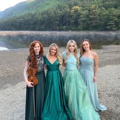 three women in long dresses standing next to each other near a body of water with mountains in the background