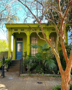 a yellow house with green trim and blue door in the front yard is surrounded by greenery