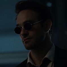 a man with round sunglasses on his face looking off to the side in a dimly lit room