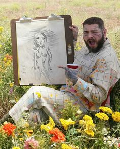 a man sitting in the grass holding a cup and drawing on an easel next to flowers