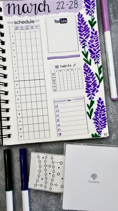 an open planner with purple flowers on it and two pens next to it in front of the calendar