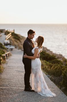 a bride and groom standing on the side of a road near the ocean at sunset