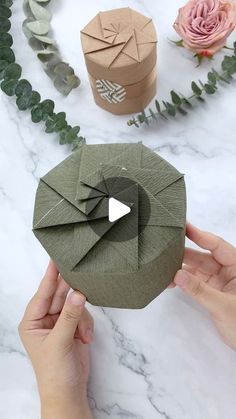 someone holding an origami box in their hands with flowers and greenery around it