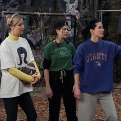 three women standing next to each other in front of a chain link fence with leaves on the ground