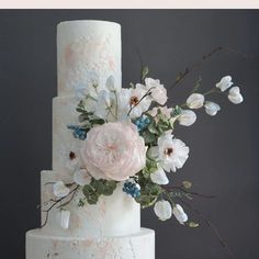 a white wedding cake with pink and blue flowers on the top, surrounded by greenery