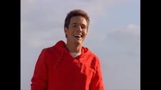 a man in a red hoodie smiles at the camera while standing under a cloudy blue sky