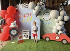 a young boy standing in front of a birthday backdrop with cars and balloons on the grass