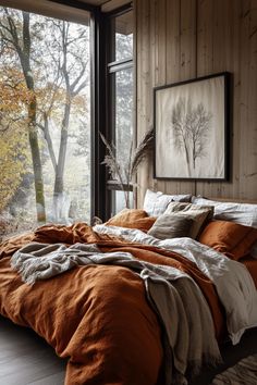 a bed with an orange comforter sitting in front of a window next to a painting