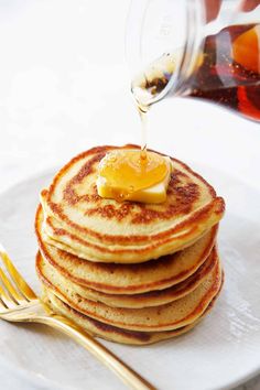 a stack of pancakes with syrup being poured onto them on a white plate next to goldware