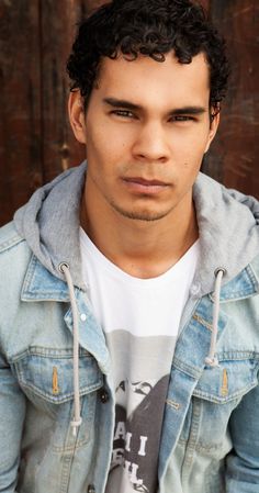 a young man with curly hair wearing a denim jacket and looking at the camera while sitting in front of a wooden wall