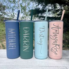 three different colored tumblers with straws in them on a wooden table near some trees