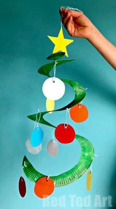 a hand holding a paper plate christmas tree decoration with ornaments hanging from it's sides