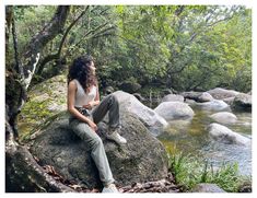 a woman sitting on top of a large rock next to a river in the forest