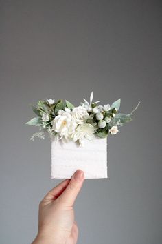 a hand holding up a piece of paper with flowers on it and greenery in the middle