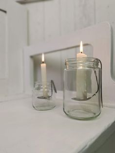 two lit candles sit in glass jars on a white countertop next to a mirror