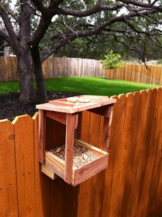 a wooden bird feeder on top of a fence next to a tree and grass area