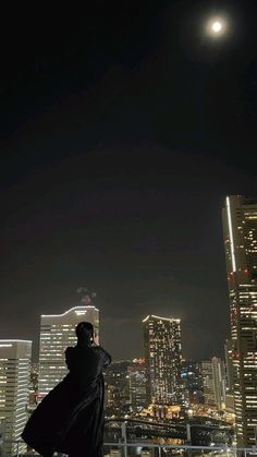 a man standing on top of a roof next to tall buildings at night with the moon in the sky