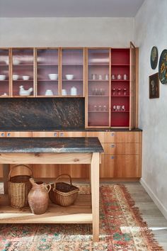 Closeup of a kitchen with a wooden work table sat on a colourful Moroccan rug in the foreground and glazed wall cabinets in the background. Red Kitchen Walls, London Plane, Glass Cabinets, Fluted Glass, Wall Cabinets, Big Kitchen