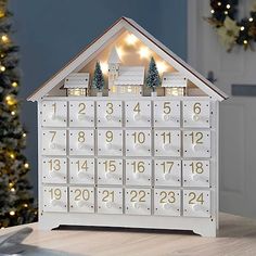 a white wooden house shaped calendar with christmas lights on the roof and trees around it