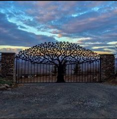 an iron gate with a tree in the middle and clouds above it at sunset or dawn