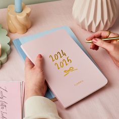 a person writing on a pink notebook with gold lettering