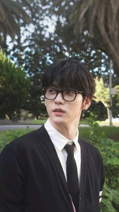 a young man wearing glasses and a black tie standing in front of some bushes with palm trees behind him