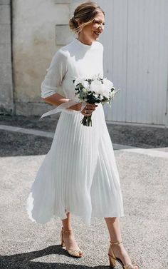 a woman in a white dress is walking down the street with flowers on her hand