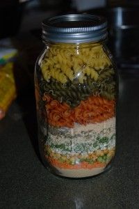 a glass jar filled with pasta and veggies on top of a counter next to corn