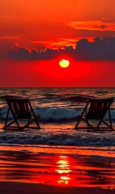 two chairs sitting on top of a sandy beach under a red sky with clouds at sunset