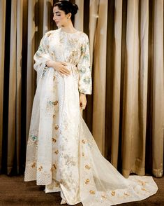 a woman standing in front of curtains wearing a white dress with gold and blue flowers on it