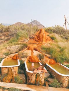 a woman laying in an outdoor bathtub surrounded by rocks and grass, with mountains in the background