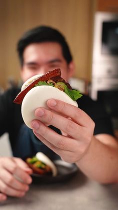 a man holding up a sandwich with bacon and lettuce on it while sitting at a table